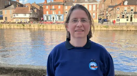 BBC/Seb Cheer A woman with brown hair wears glasses and a sweatshirt reading "York Rescue Boat". She is standing on Queens Staith in York with the River Ouse in the background.