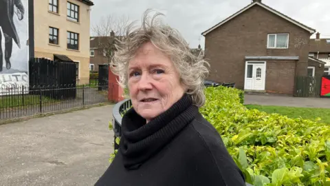 Carmel McCallion. She has grey-ish hair, looking sideways at the camera and wearing a black roll-neck jumper. Houses can she seen in the background.