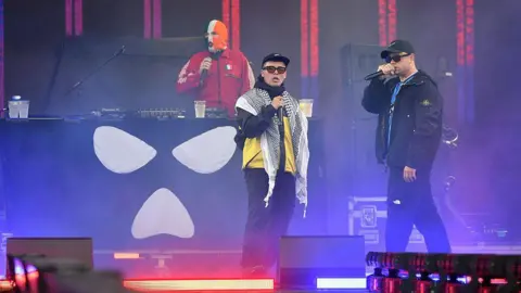 Two rappers perform on stage with microphones, wearing sunglasses and streetwear, while a masked DJ stands behind a booth decorated with a white skull-like symbol. Purple and blue lights fill the stage.