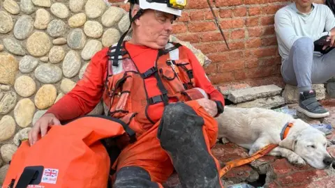Essex County Fire & Rescue Service A man is slumped sitting down on bricks and rubble, with a bright orange rescue outfit on. A dog is lying down with its eyes closed next to him.