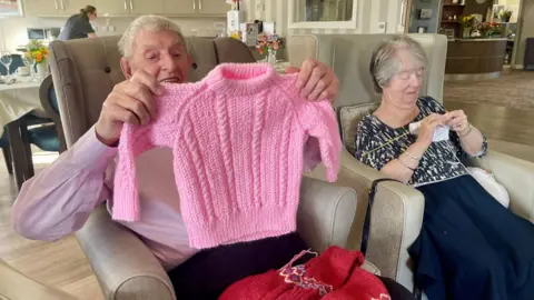 A man with grey hair sits in an armchair holding up a small pink woollen jumper. Another maroon knitted jumper sits on his lap. The man is wearing a pink shirt. A lady sits beside him also in a grey armchair. She is knitting using white wool. She is wearing a black and white patterned blouse with a dark skirt. Behind them, dining tables are set with crockery and glasses. A woman in a black uniform and glasses can be seen beside some overhead cabinets.