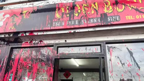 A Chinese takeaway splattered with red and black paint. The paint covers the red sign and the the two glass windows either side of the glass door. The sign reads The Golden Bowl in yellow lettering.
