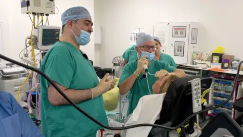 The picture shows a hospital theatre, two men with green scrubs and blue net hats are treating a woman on a hospital bed. They are wearing latex gloves and blue face masks. The woman is sitting up-right and is facing away from the camera.