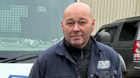 Gary Hallett has a short beard and is wearing black zip up top and blue overalls. He is stood in front of a white van and is looking at the camera.