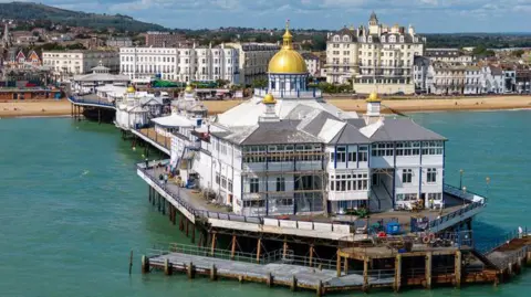 An aerial view of Eastbourne Pier shows a large white pavilion building at the end featuring a central gold dome and smaller domes. The structure stands on dark metal supports above the sea, with wooden walkways and railings visible around its perimeter. Behind the pier, seafront hotels and apartments line a sandy beach, with hills in the background.