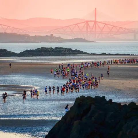 Pauline Bell a large group of people all wearing bright colours running across a beach. the forth rail bridge is visble in the distance,