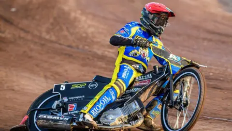 Getty Images Tai Woffinden races his bike on a shale speedway track. He is wearing a red helmet and blue and yellow race suit. The motorbike is leaning and sliding around a corner with a spray of dirt visible from the back wheel.