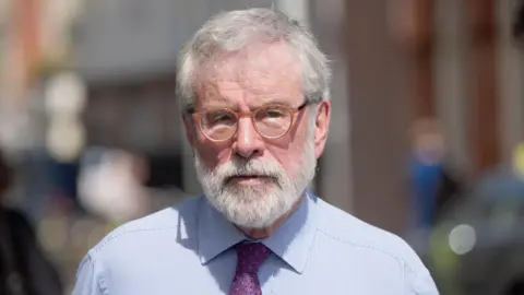 Gerry Adams walks outside the High Court in Dublin. He has short white hair and beard. He is wearing small, orange circular glasses and his expression is neutral. He is also wearing a light blue collared shirt and a purple patterned tie.

