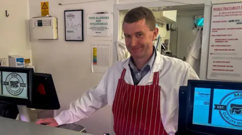 BBC/ Emily Johnson A butcher stands behind the counter in a traditional butcher shop, wearing a white coat and a red-and-white striped apron. Two digital screens on the counter display the logo “M&K Quality Butchers EST. 1970”