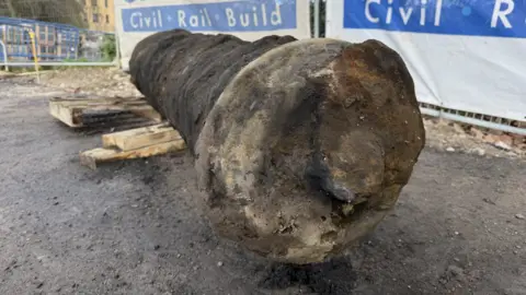 BBC / Becki Bowden The photo shows a cast iron metal bar which has been laid down. The face has gone rusty. It has been laid on a wooden pallet. There are white and blue banners on the fences, reading 'Civil, Rail, Build.' 