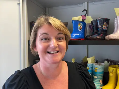 Laura Tommy has short, light brown hair and is wearing a black top. She is standing in front of some shelves which have an array of children's wellies on them.