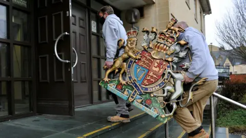 Two workers carry a royal coat of arms shield walking up the steps into the courthouse, while wearing face masks