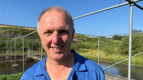 A man with short grey hair smiling at the camera. He is wearing a blue polo-style shirt. Behind him is netting running over large scaffolding structure and hills