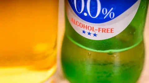 A pint glass which is full of beer, and a green beer bottle. The beer bottle has a sticker on it which reads "0.0% Alcohol-Free".