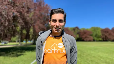 Dr Hussain Al-Zubaidi stands in War Memorial Park in Coventry. Behind him is a vivid blue sky, trees and bright green grass. He has black short hair and is wearing an orange parkrun top and a grey sports jacket over the top.
