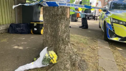 Mousumi Bakshi/BBC A bouquet of flowers at the base of a tree, with a police vehicle to the right. A number of people are standing around in the distance. There are bags to the left and a large tree trunk with police tape around it. 