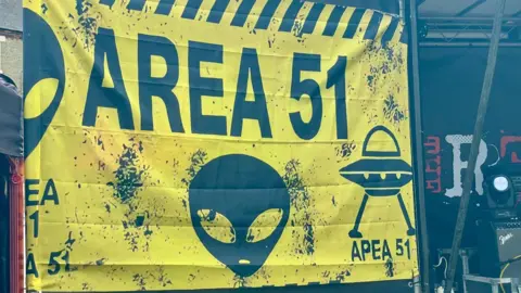 Stephen Huntley/BBC A yellow sign has "AREA 51" written on it. There are pictures of an alien's head and a UFO, alongside splattered paint marks.