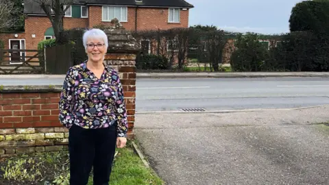 Alison Thomas is standing in her front garden and next to a driveway. Behind her we can see a single carriageway road. She's smiling and wearing glasses and a multi-coloured long-sleeve top.