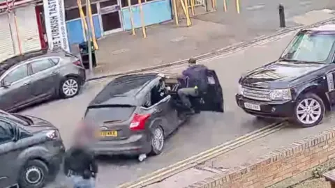 A man his hanging on to a car while half portruding from the passenger side door. The car is pulling away