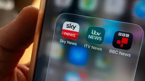Hand holding a phone, that shows ITV and Sky and BBC news apps on the phone screen.