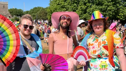 Three attendees smile at the camera. All wearing colourful outfits and holding fans in the heat. Two have facepaint, two have hats, including a rainbow one