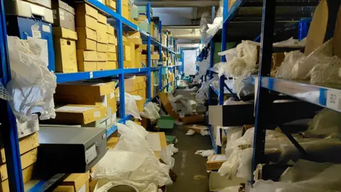 Bristol City Council An interior shot of the Bristol Museum store. There are two long rows of blue metal shelves receding into the distance with manila coloured storage boxes on the shelves. Many of the boxes have been pulled onto the floor and are half-open, with lids, packaging and some antiques scattered on the floor.