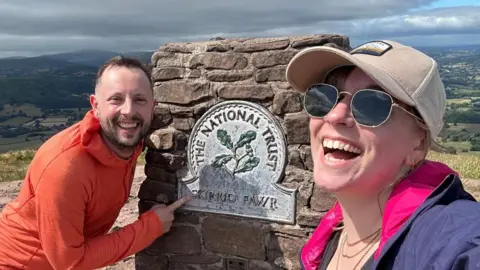 Jody and Jessie stand on Skirrid Fawr walk. Jody is wearing an orange half zip jacket. Jessie is taking a selfie and wearing a purple jacket, white hat and sunglasses. 