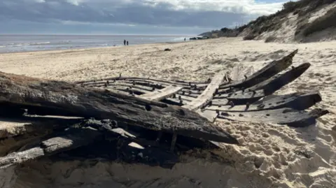 Shaun Whitmore/BBC The old timbers lie in the foreground. Beyond them, the beach - with low sandy cliffs on the right - stretches into the distance. Two tiny figures can be seen on the shoreline.