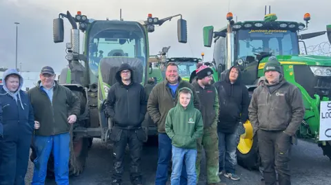 Nine men stand in front of two green tractors. Some of the tractors have signs that say: "Support local produce this Christmas".