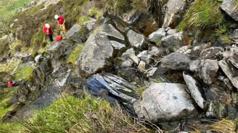 North Wales Police A dark blue jacket is just visible by a pile of rocks. Below, two mountain rescue members wearing white helmets and red t-shirts are examining the ground.