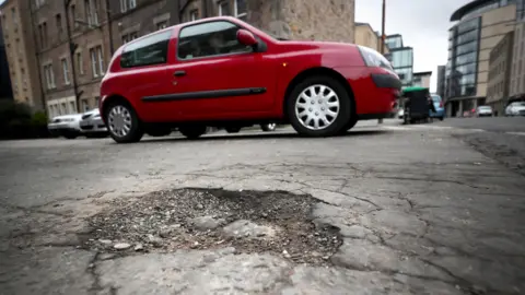 A red car drives past an unfilled pothole on a street