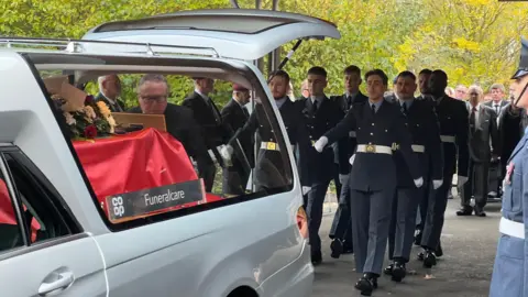 Servicemen in navy uniforms with white belts and white gloves march towards an open hearse. The coffin is draped in a red flag with a large floral arrangement on top. 