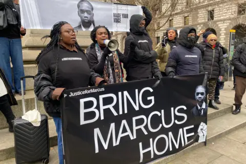 About 10 campaigners holding a large black banner, with white lettering saying BRING MARCUS HOME. One person has a megaphone and they are wearing tops with 'justiceforMarcus' on.