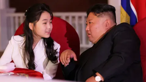 Reuters Kim Ju Ae (left) and Kim Jong Un (right) speak to each other. They are sitting at a table on red chairs. She is looking at him