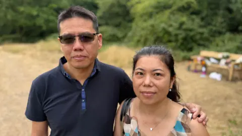 Franky Lau and Jessie Deng look towards the camera while standing in front of a wooden bench commemorating their daughter Selena.