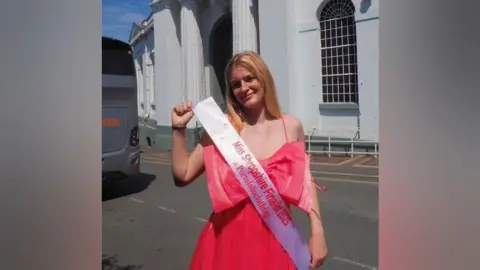 Nell Massin A woman with strawberry blonde hair is wearing a pink dress with a bow on the front. She is wearing a sash that reads "miss shropshire finalist 2025"