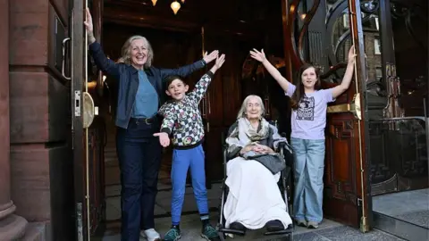 Greg Macvean Photography Four people in the open doorway of a theatre - a woman, a boy and a girl standing with their arms held aloft, and an older woman sitting in a wheelchair smiling.
