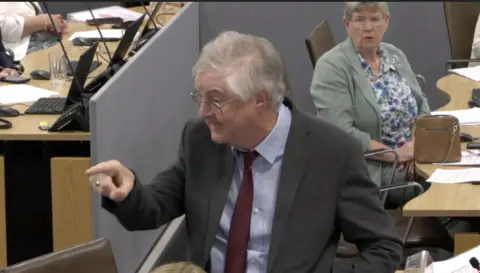 Mark Drakeford who has grey hair and glasses, wearing a grey suit jacket, shirt and red tie, pointing his finger and looking to the side with an angry expression. Behind him, sat nehind a wooden Senedd desk, is colleague Jan hut who also has short grey hair and glasses and is wearing a mint green blazer and blue and white floral blouse. She is looking at Drakeford with a concerned expression.