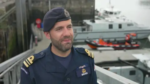 A picture of Lieutenant Commander Ben Brown. He is smiling, while stood in front of a number of boats.