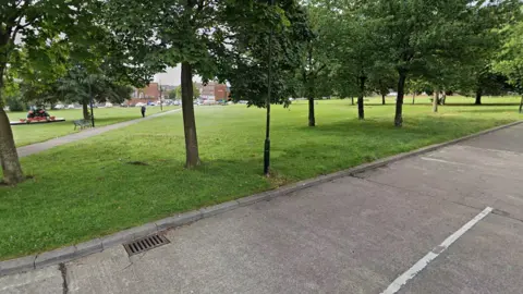 Google A wide shot of a grassy square ringed with trees with a path going through the centre of it
