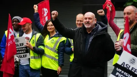 Victoria Jones/PA Media Bearded man wearing large blue coat stands with one arm raised among strikers wearing yellow hi-viz