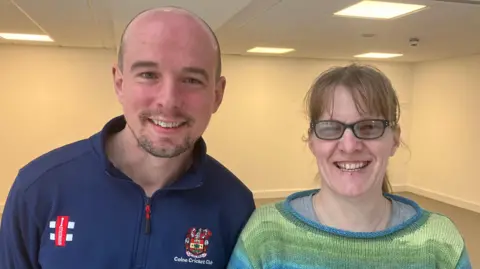 A man and a woman smile in front of a simple interior background. The man on the left is bald with stubble and is wearing a top that reads Colne Cricket Club. The woman has strawberry blonde hair, glasses, and a green and blue jumper