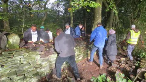 Bradford Council A group of people, one in a high vis jacket, on either side of a damaged dry stone wall. They are all working on part of the wall. Trees behind them in the background.