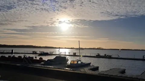 Two boats are moored up at a pontoon in the foreground of the picture. Behind them an expanse of water stretches to the horizon. The sky is still blue but darkening with a hazy setting sun in its centre.