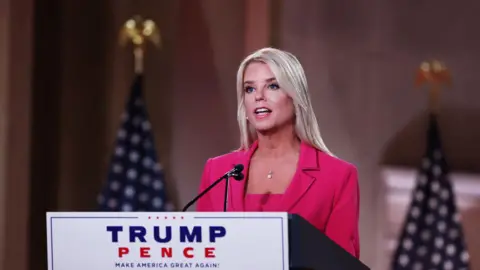 Pam Bondi, standing before a lectern and speaking into a microphone. She is wearing a pink suit. 