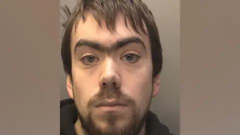 A mugshot of Ryan Woosley with a brown beard, short hair and a monobrow