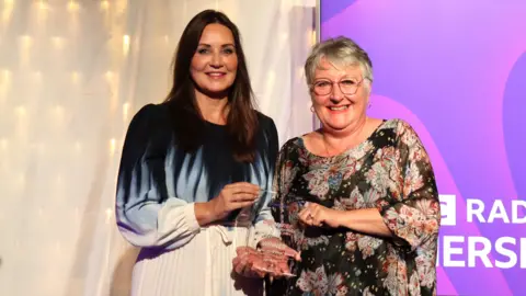 Natalie Dyson and Lyn Foley on stage, each holding a corner of Ms Foley's glass award.