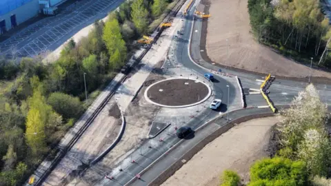 An aerial view of a new roundabout, which has dirt in the middle and white concrete around the circumference. Three partially completed roads link to the roundabout, with work still going on. Trees are visible on either side.