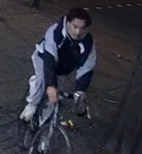 Avon & Somerset Police A grainy CCTV image of a young man in a black and grey tracksuit, riding a bike at nighttime. 