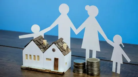 A model of a house sits next to a pile of coins and a paper cut out of a family. It is all placed against a blue background. 